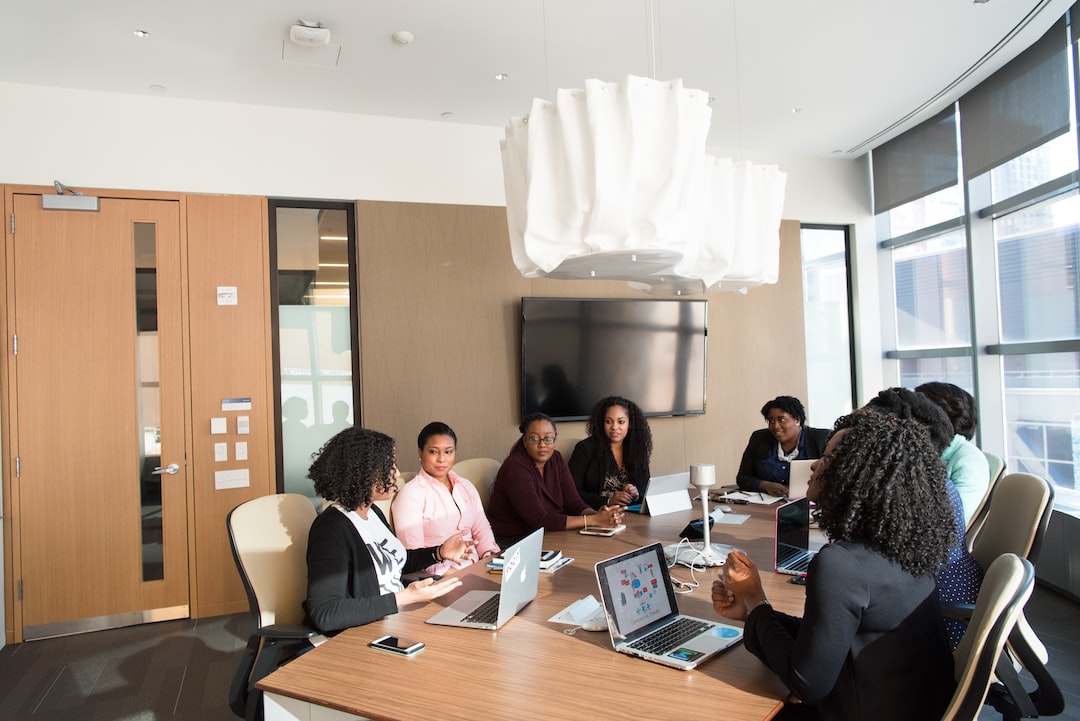 A diverse team of professional women engaged in a business meeting around a conference table with laptops in a bright modern office.